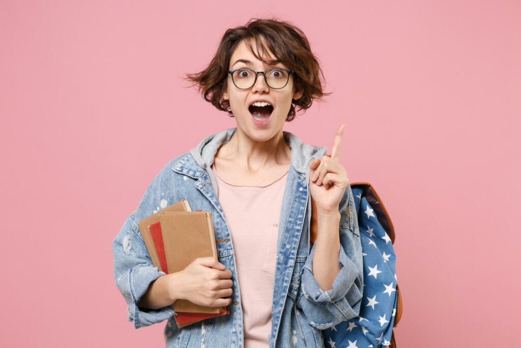 Excited girl student in denim clothes glasses backpack isolated on pastel pink background. Education in high school university college concept. Hold books pointing index finger up with great new idea