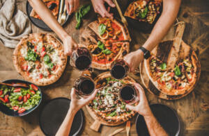 Family or friends having pizza party dinner. Flat-lay of people clinking glasses with red wine over rustic wooden table with various kinds of Italian pizza, top view. Fast food lunch, celebration