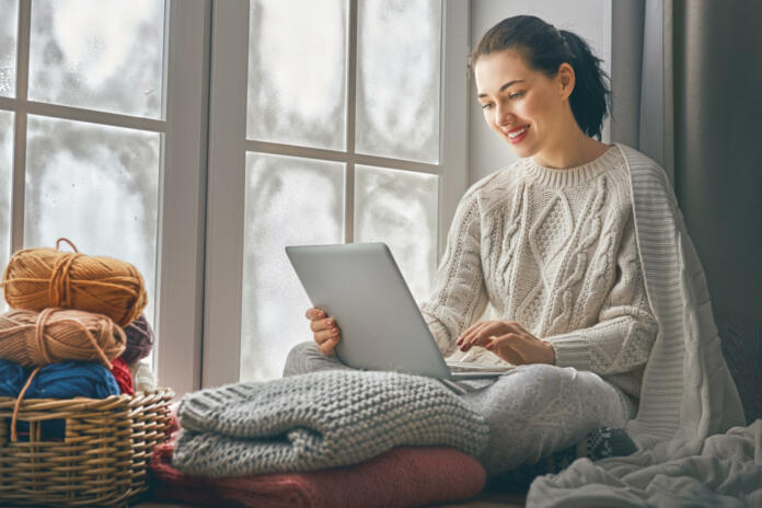 Happy beautiful woman watching videos or enjoying entertainment content on a laptop sitting near windows in the house in the winter.