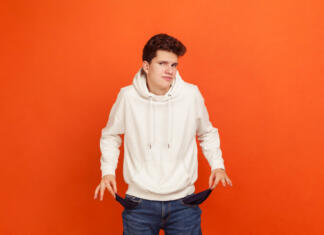 Svetovalnica – vračanje štipendije? I have no money, Poor teenager in casual sweatshirt with hood demonstrating empty pockets of his modern denim pants, low state stipend, bankruptcy. Indoor studio shot isolated on orange background