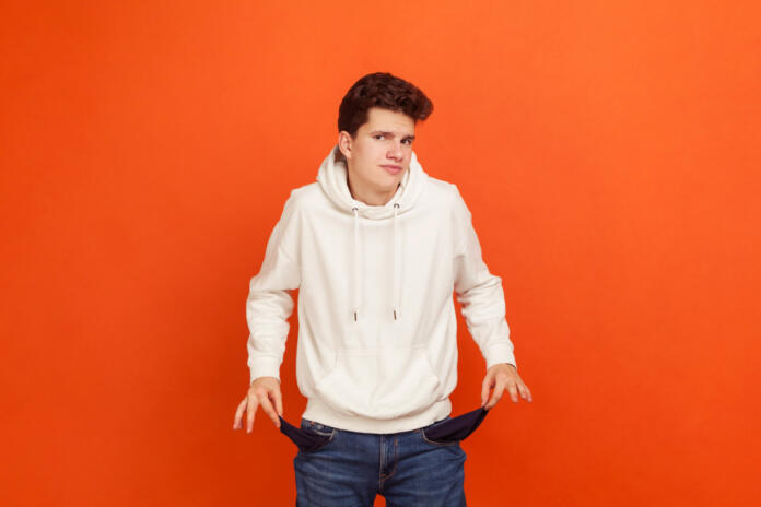 I have no money, Poor teenager in casual sweatshirt with hood demonstrating empty pockets of his modern denim pants, low state stipend, bankruptcy. Indoor studio shot isolated on orange background