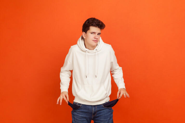 I have no money, Poor teenager in casual sweatshirt with hood demonstrating empty pockets of his modern denim pants, low state stipend, bankruptcy. Indoor studio shot isolated on orange background