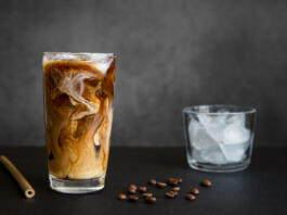 Iced coffee in tall glass with cream, container with ice, cocktail straw and coffee beans on dark background with copy space. Refreshing drink.