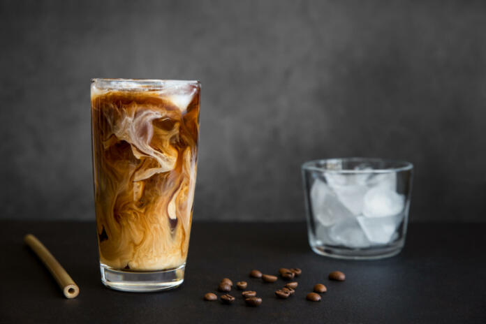 Iced coffee in tall glass with cream, container with ice, cocktail straw and coffee beans on dark background with copy space. Refreshing drink.