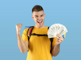 Planning travel budget. Funky millennial hipster with tourist gears and fan of dollars gesturing YES over blue studio background. Excited young guy having enough money for his dream journey