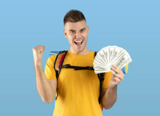 Akontacija na napotnico po 26. letu starosti Planning travel budget. Funky millennial hipster with tourist gears and fan of dollars gesturing YES over blue studio background. Excited young guy having enough money for his dream journey