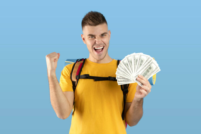 Planning travel budget. Funky millennial hipster with tourist gears and fan of dollars gesturing YES over blue studio background. Excited young guy having enough money for his dream journey