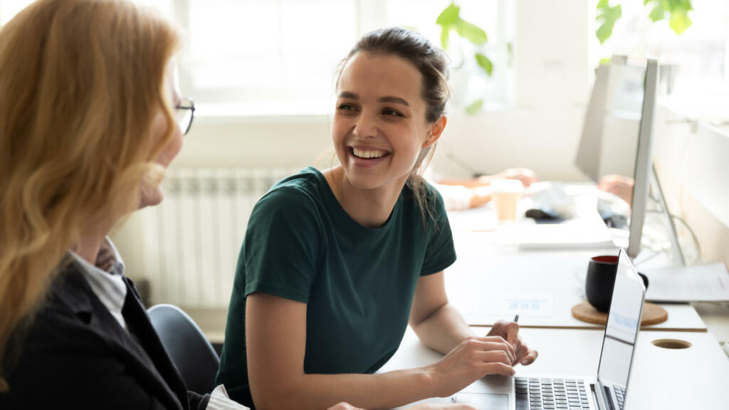 Pleasant workday. Two women colleagues employees of diverse age sharing points of view at workplace in office, happy smiling millennial female trainee discussing business ideas with adult aged trainer