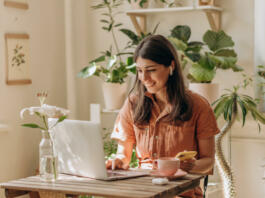 Positive young mixed race woman using a laptop and smartphone at home.Cozy home interior with indoor plants.Remote work, business,freelance,online shopping,e-learning,urban jungle concept.