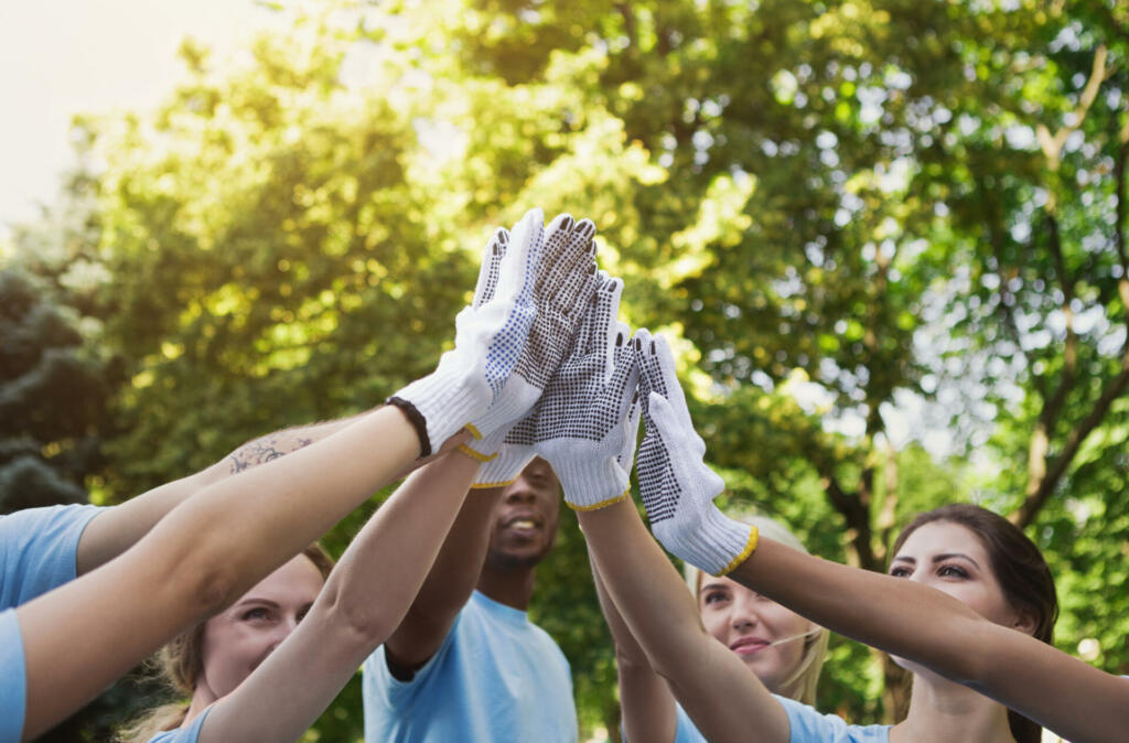 Volunteering, unity and environment care concept. Group of young volunteers in protective gloves making high five before cleaning park, closeup