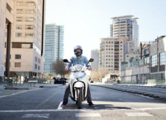 Mladi v cestnem prometu: Priročnik projekta BE A HERO Wide shot of a young motorcyclist stopped at a traffic light in Barcelona. The man riding his scooter through the city on a large avenue lined with skyscrapers is waiting at the traffic light.