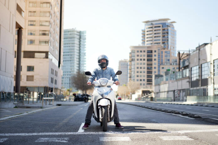 Wide shot of a young motorcyclist stopped at a traffic light in Barcelona. The man riding his scooter through the city on a large avenue lined with skyscrapers is waiting at the traffic light.