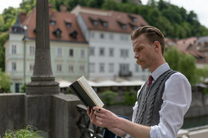 A young hipster man with a mustache on a walk around the city drinking coffee,  reading a book .