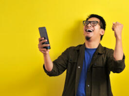 Asian man holding smartphone with winning gesture. Asian bussinesman winning gift or lottery, on yellow background