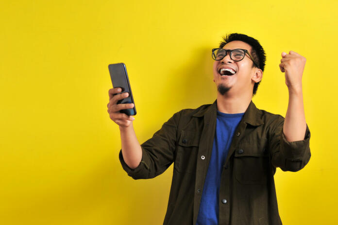 Asian man holding smartphone with winning gesture. Asian bussinesman winning gift or lottery, on yellow background