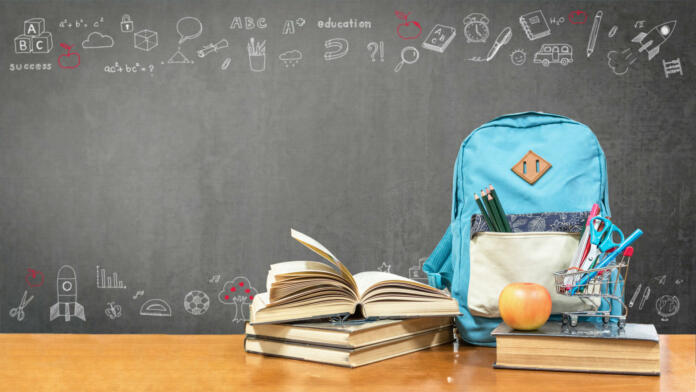 Back to school concept with school books, textbooks, backpack and stationery supplies on classroom desk with teacher's black chalkboard background with educational doodle for new academic year begin