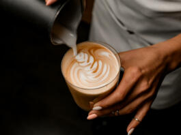 Close-up of barista gently drawing pattern pouring milk from jug into glass with latte in her hand