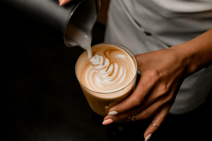 Close-up of barista gently drawing pattern pouring milk from jug into glass with latte in her hand