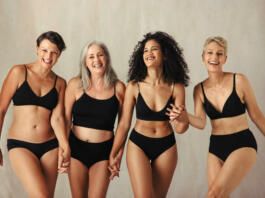 Female models of different ages celebrating natural bodies. Four body positive and confident women smiling cheerfully while wearing black underwear and holding hands together in a studio.