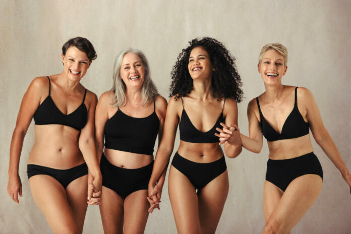 Female models of different ages celebrating natural bodies. Four body positive and confident women smiling cheerfully while wearing black underwear and holding hands together in a studio.