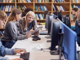 Female University Or College Student Working At Computer In Library Being Helped By Tutor