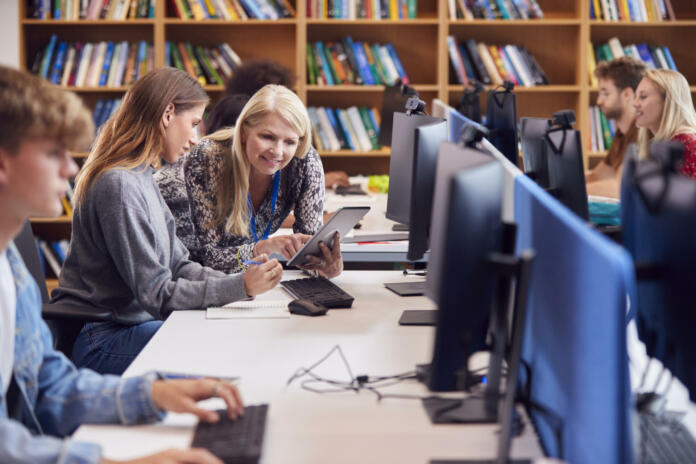 Female University Or College Student Working At Computer In Library Being Helped By Tutor