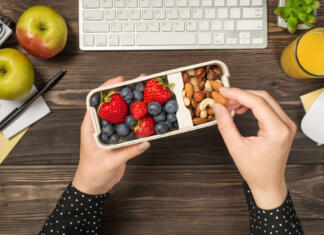 Dieta za nadpovprečno zdravje First person top view photo of woman's hands holding lunchbox with healthy meal nuts and berries over apples glass of juice flowerpot stationery keyboard mouse on isolated dark wooden table background