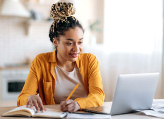 Študentsko delo in novosti Focused cute stylish african american female student with afro dreadlocks, studying remotely from home, using a laptop, taking notes on notepad during online lesson, e-learning concept, smiling