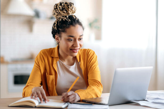 focused-cute-stylish-african-american-female-student-with-afro-dreadlocks-studying-remotely-from-home-using-a-laptop-taking-notes-on-notepad-during-online-lesson-e-learning-concept-smiling-stockpack-istock Focused cute stylish african american female student with afro dreadlocks, studying remotely from home, using a laptop, taking notes on notepad during online lesson, e-learning concept, smiling