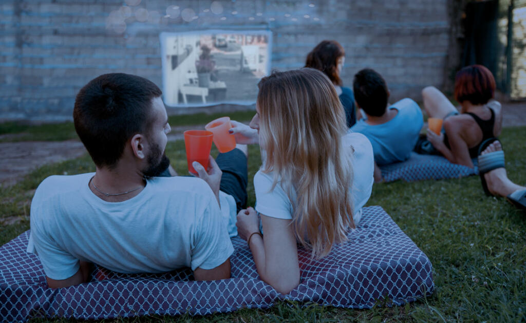 Friends laying cozy in green grass and watching film at open cinema in Home backyard. Perfect spending weekend time with family