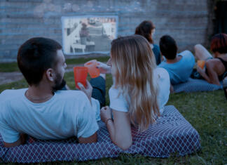 Poletni filmski biseri: 12 čudovito poletnih filmov Friends laying cozy in green grass and watching film at open cinema in Home backyard. Perfect spending weekend time with family