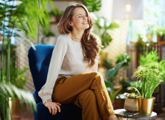 Šola za prave ženske Green Home. smiling elegant woman with long wavy hair at modern home in sunny day in green pants and grey blouse sitting in a blue armchair.