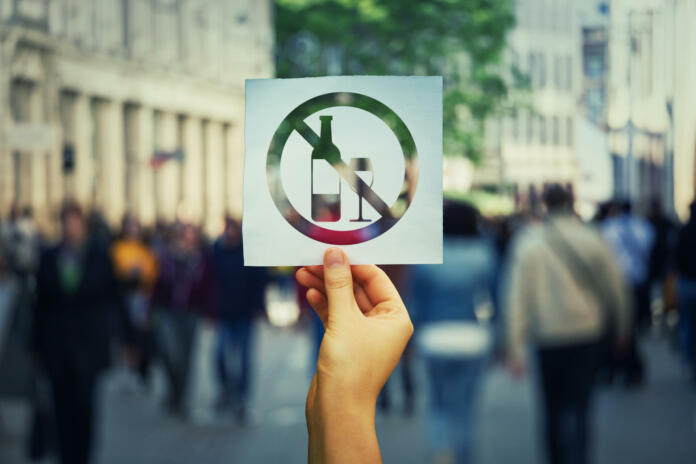 Hand holding a paper sheet with no alcohol sign over a crowded street background. Stop to drink symbol prohibited icon. Refuse to be dependent.