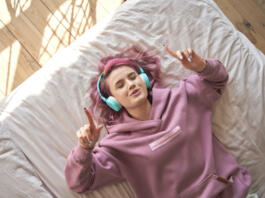 Happy funny teen girl with pink hair wear headphones lying in comfortable bed listening new pop music enjoying singing song with eyes closed relaxing in cozy bedroom at home. Top view from above.