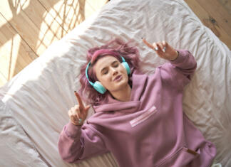 Grem v študentski dom! Happy funny teen girl with pink hair wear headphones lying in comfortable bed listening new pop music enjoying singing song with eyes closed relaxing in cozy bedroom at home. Top view from above.