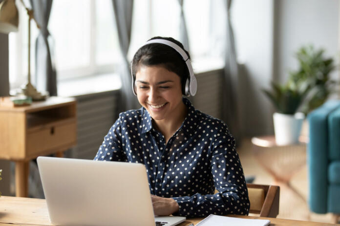 Happy indian young girl student wear headphone watch webinar listen online course communicate by conference video call e learn language in app laugh study with teacher lesson look at laptop at home