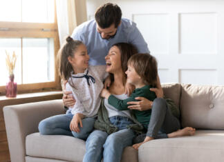 Vzdrževani in nevzdrževani družinski člani Happy young Caucasian family renters with two small kids sit rest on couch in cozy living room. Smiling parents with little children relax on sofa enjoy weekend in new renovated design home together.
