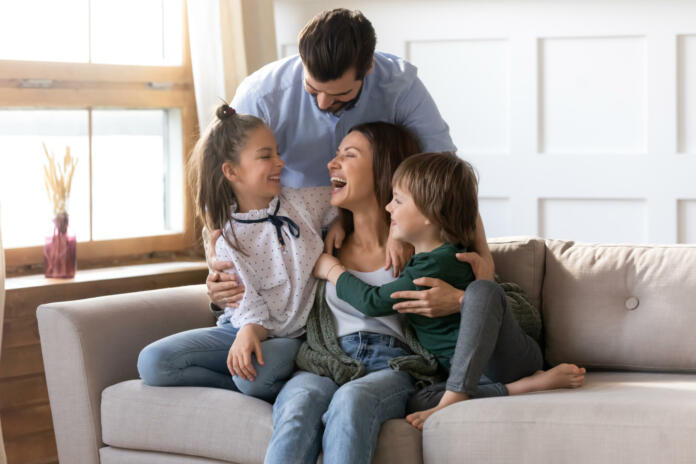 Happy young Caucasian family renters with two small kids sit rest on couch in cozy living room. Smiling parents with little children relax on sofa enjoy weekend in new renovated design home together.