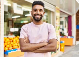 Iz moške perspektive Hispanic man standing by fruits and vegetables shop. Smiling happy with crossed arms by marketplace
