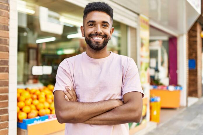 Hispanic man standing by fruits and vegetables shop. Smiling happy with crossed arms by marketplace