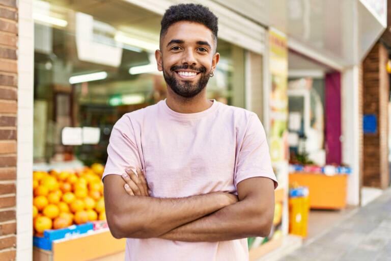 Hispanic man standing by fruits and vegetables shop. Smiling happy with crossed arms by marketplace