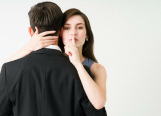 Prepovedan sadež I have a secret. Portrait of a young girlfriend making eye contact and putting a finger on her mouth while hugging her boyfriend or husband
