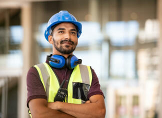 Najbolj iskani poklici Indian construction site manager standing with folded arms wearing safety vest and helmet, thinking at construction site. Young architect watching construction site with confidence. Portrait of mixed race manual worker looks at his work with satisfaction.