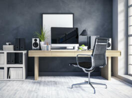 Interior of a modern home office next to a big window. Light wood desk with organized set up of books, office supplies and computer with some room decor around it.