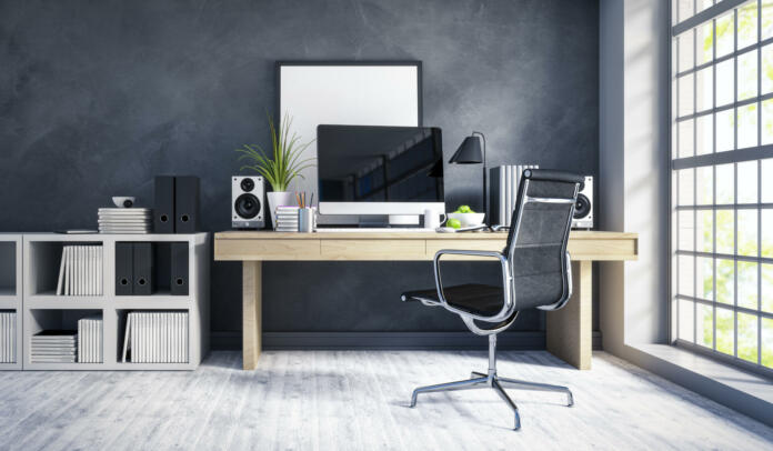 Interior of a modern home office next to a big window. Light wood desk with organized set up of books, office supplies and computer with some room decor around it.