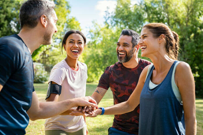 Laughing mature and multiethnic sports people putting hands together at park. Happy group of men and beautiful women smiling and stacking hands outdoor after fitness training. Multiethnic sweaty team cheering after intense training.