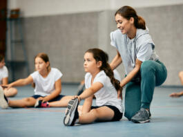 Little girl stretching on the floor and warming up with help of PE teacher during a class at school gym.