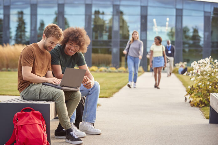 Male University Or College Students Sitting Outdoors On Campus Talking And Working On Laptop