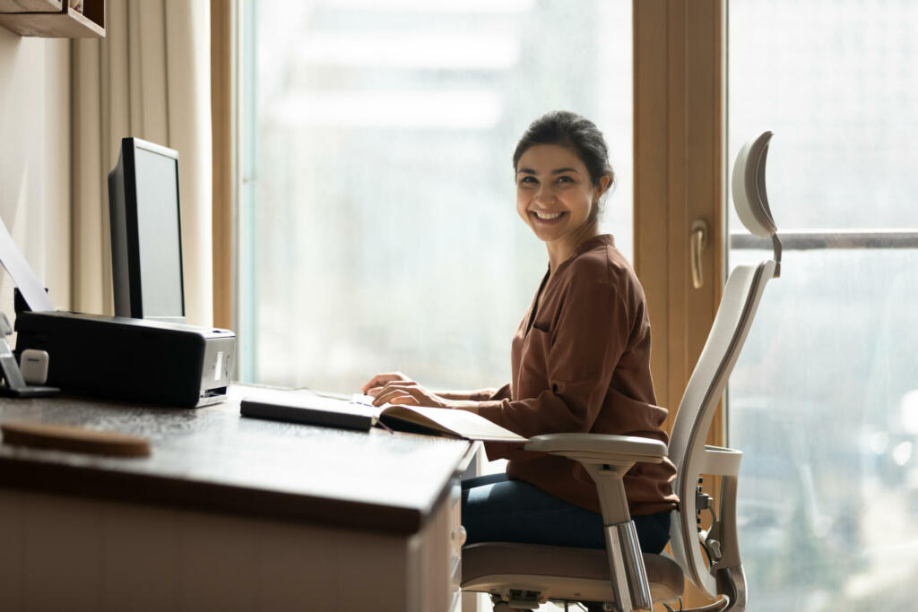 Modern day worker. Portrait of happy biracial business woman freelancer sit by computer at comfy workplace at corporate workspace or at home. Smiling young indian lady office employee look at camera
