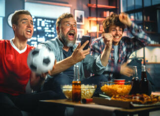 Športni komentatorji in solze smeha (drugi del) Night at Home: Three Soccer Fans Sitting on a Couch Watch Game on TV, Use Smartphone App to Online Bet, Celebrate Victory when Sports Team Wins. Friends Cheer Eat Snacks, Watch Football Play.
